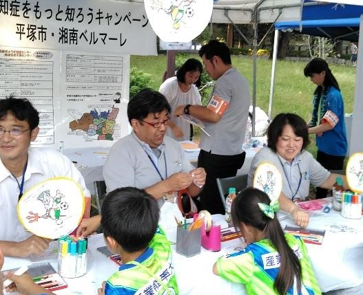 小学生が、キングベルに塗り絵をして、うちわに貼りつけるイベントが行われました。平塚市地域包括ケア推進課職員、平塚市高齢者よろず相談センター職員とともに。