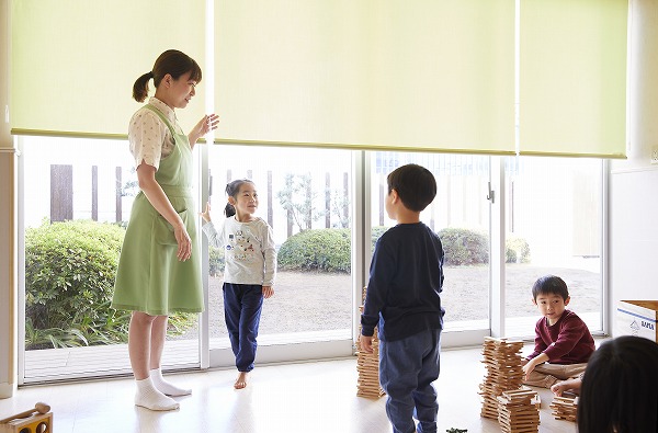 ロールスクリーンを操作する松村先生と室内で遊ぶ子どもたち