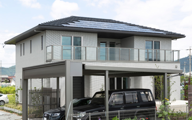 パナホーム愛岐｜モダンなデザインの二階建て住宅。バルコニーと駐車スペースには車が停まっており、快適なライフスタイルがうかがえる。