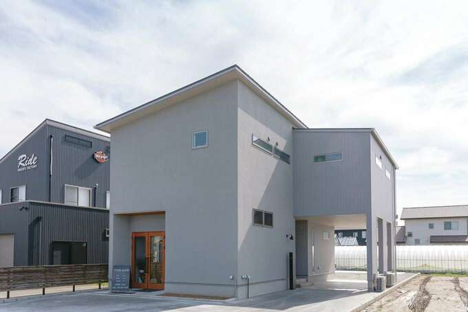 淡いグレーを基調にした外観 正面は塗り壁にして木扉を付け 店舗の入り口に 縦長の敷地を有効活用し 西面にピロティ付きの自宅用玄関を設置 奥に