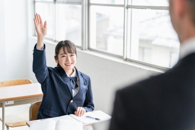 明るい教室で、先生が笑顔で生徒に話しかけ、生徒が手を挙げて積極的に発言している様子。性の多様性について安心して話し合える教育現場の雰囲気を表現