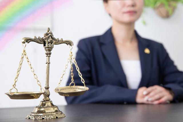 弁護士バッジを付けた女性弁護士が机に座り、金色の天秤を前に手を重ねている構図。同性婚訴訟と婚姻の自由を象徴するイメージのサムネイル