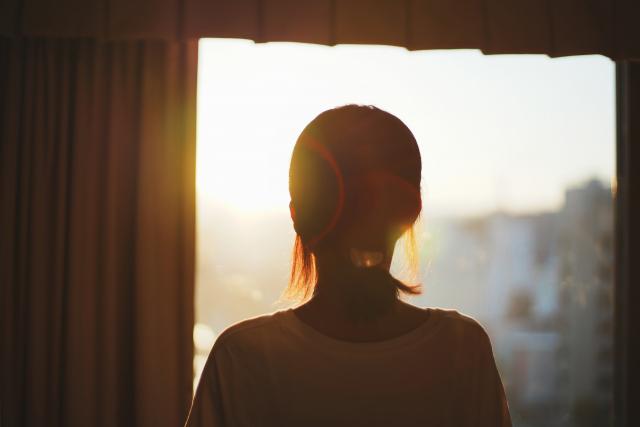 夕暮れの光が差し込む窓辺で外を見つめる女性のシルエット。カミングアウトの葛藤や家族との再生を象徴する静かな情景。