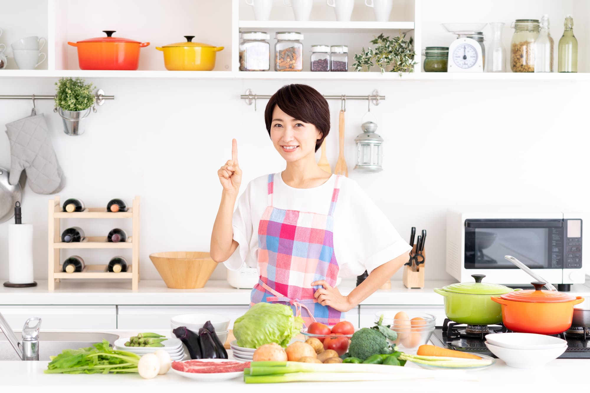 食材がたくさん並んだキッチンで、人さし指を上に向けて笑顔を浮かべる女性