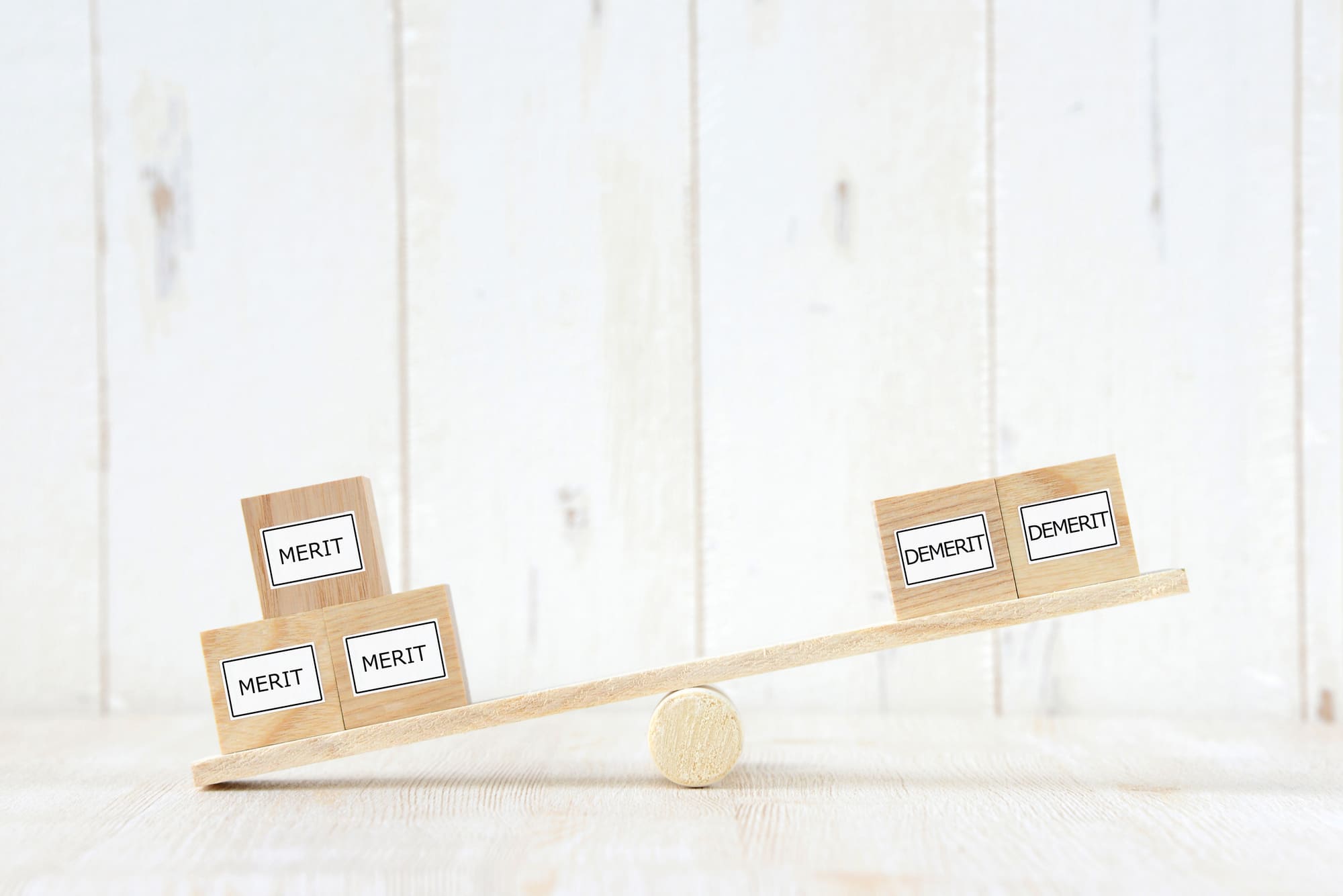 シーソーの上に乗った「MERIT」と「DEMERIT」の木のブロック