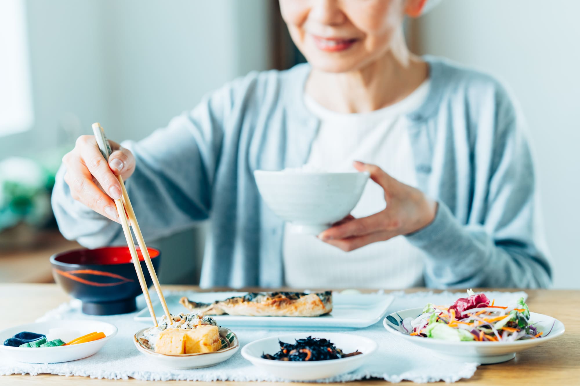 たくさんの皿が並ぶ食卓で、玉子焼きに箸を伸ばすシニア女性