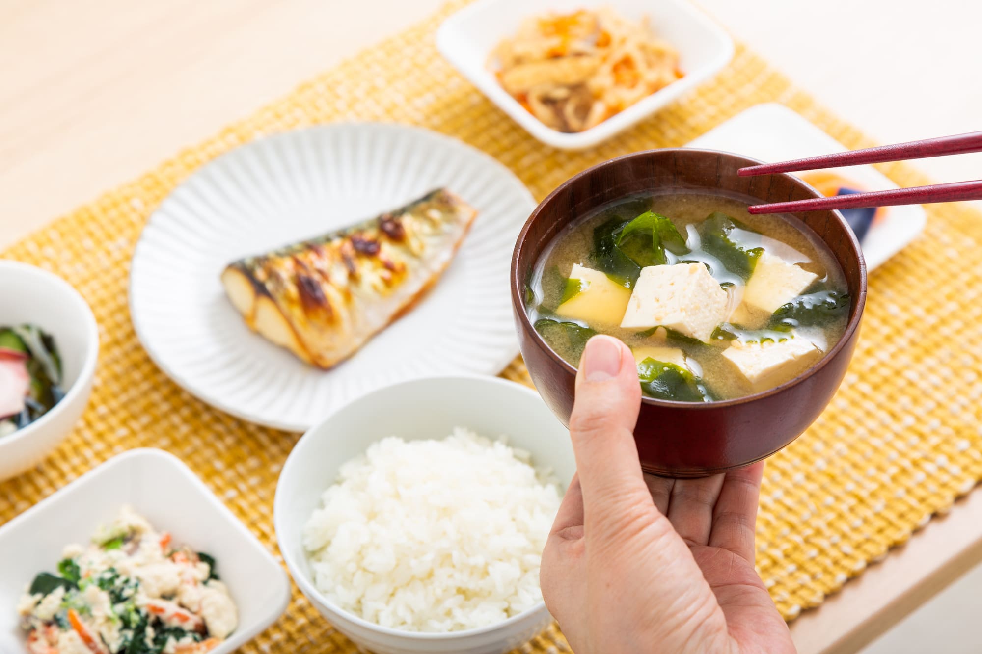 食卓に並べられた和食と、豆腐とわかめが入ったみそ汁を食べようとする人の手