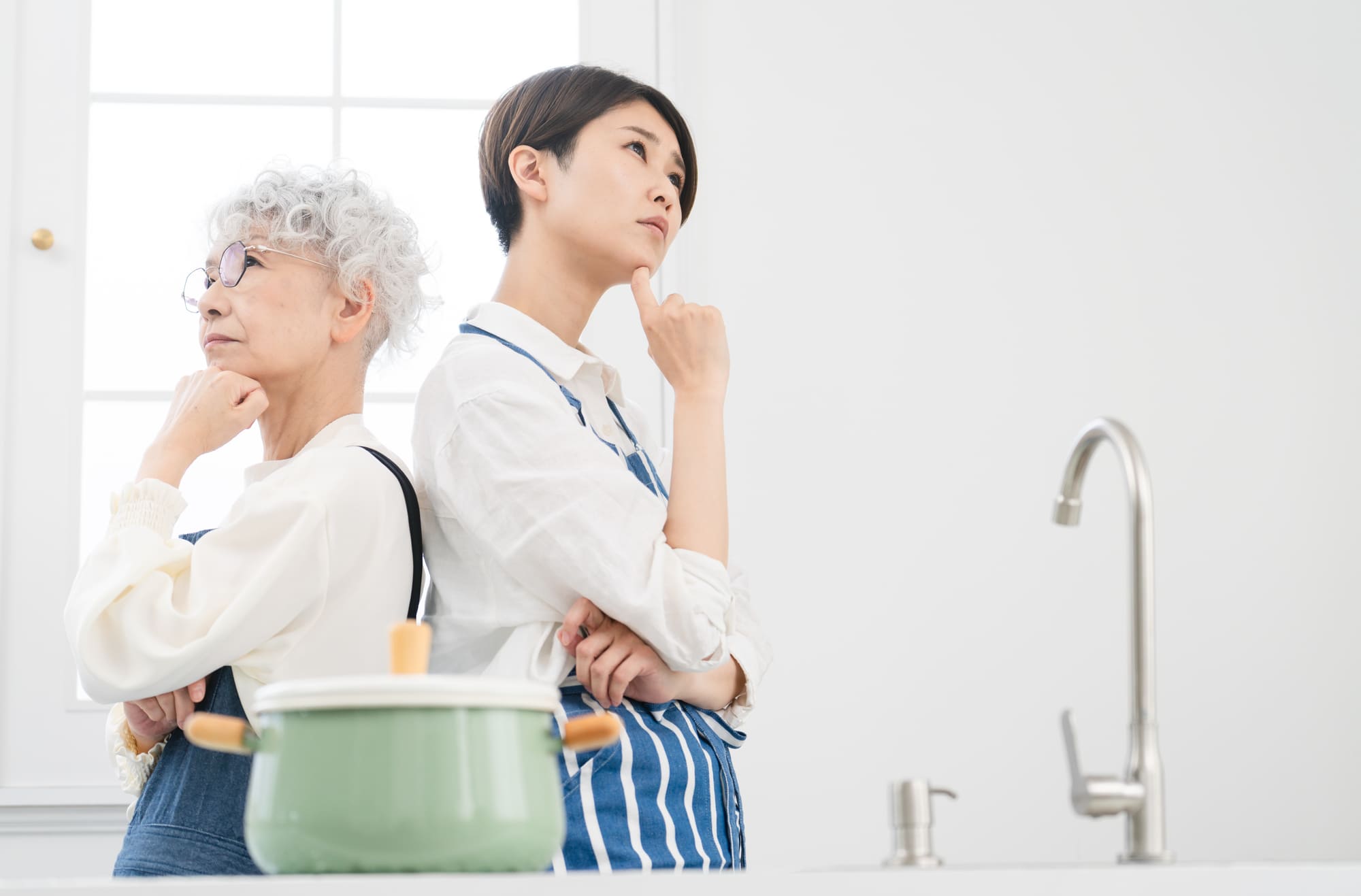 キッチンで顎に手を当てて悩んでいる若い女性とシニア女性