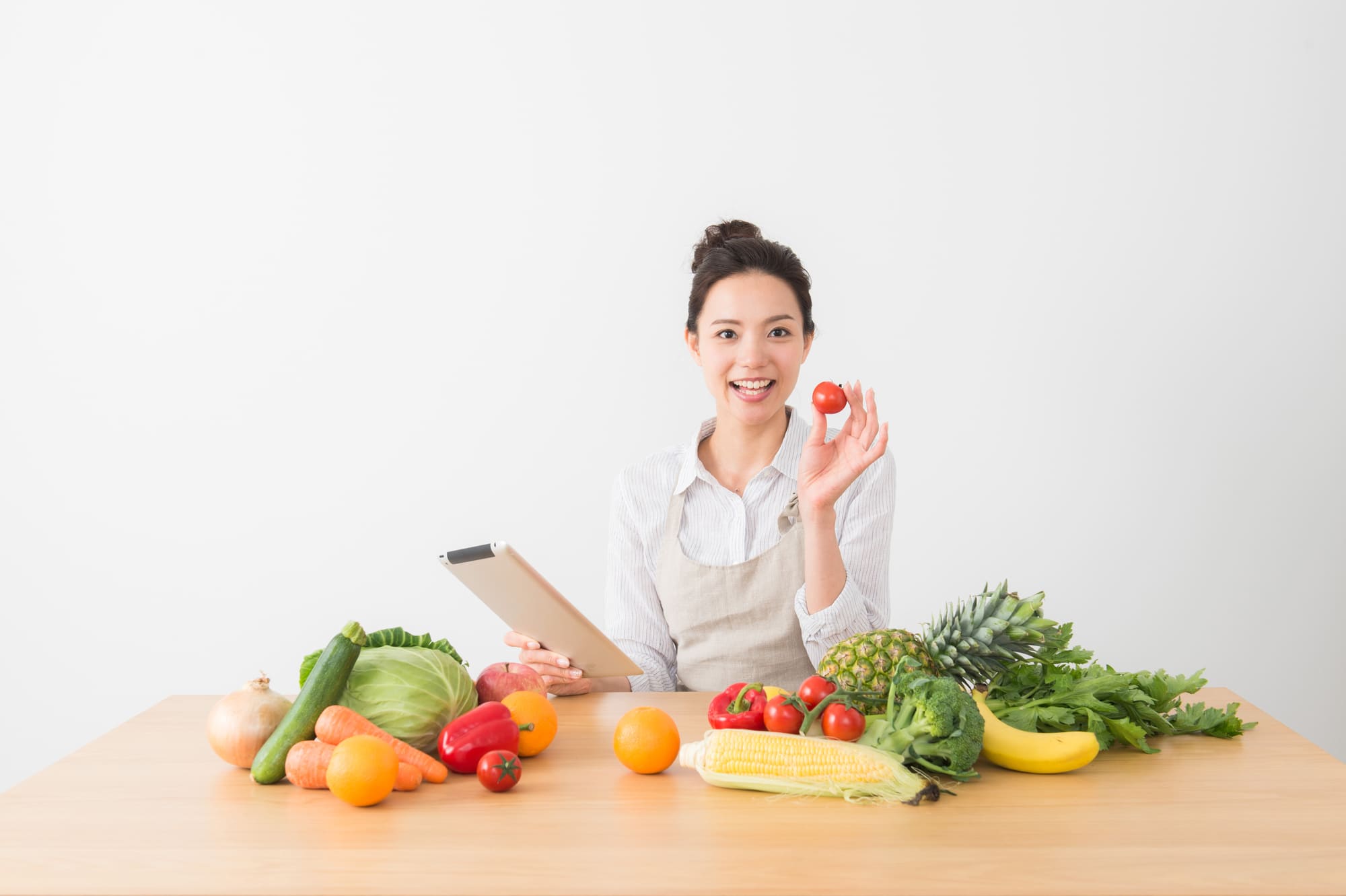 机の上に並んださまざまな食材、トマトを手に持った笑顔の女性