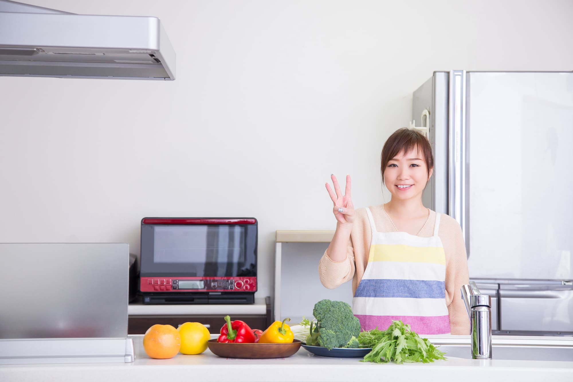 野菜が置かれたキッチンで指を3本立てるエプロン姿の女性