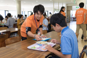 ご飯を載せたトレイを良く見てみると、カラフルなトレーマットが。実はこのトレーマットには、酪農大クイズが10問載っています。ご飯を食べ終わった高校生の皆さんに、学生スタッフがお声掛け。ここで皆さんにもクイズをひとつ。「Q、白黒のホルスタイン、毛を刈ると地肌の色は何色でしょう?」A、毛と同じく白黒 B、真っ白 C、真っ黒 皆さんわかりますか?[/