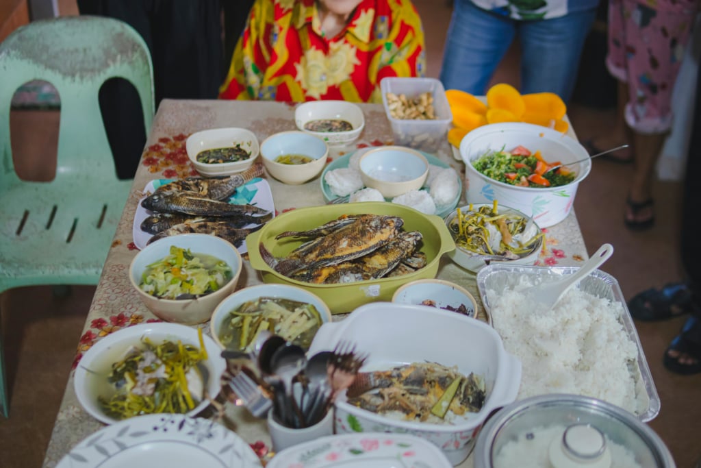 A top-down shot shows a table laden with a variety of Filipino dishes, suggesting a communal meal or feast. Several platters and bowls hold cooked fish, some appearing fried or grilled. There are also various vegetable dishes, some in broth, and a large container of steamed white rice. Other bowls contain smaller portions of what looks like dipping sauces or side dishes, and a clear container holds peanuts or similar snacks.