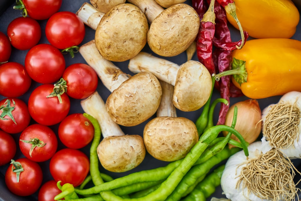 A top-down view of fresh produce including red cherry tomatoes, various mushrooms, yellow bell peppers, dried red chili peppers, green chilies, and garlic bulbs on a dark surface.