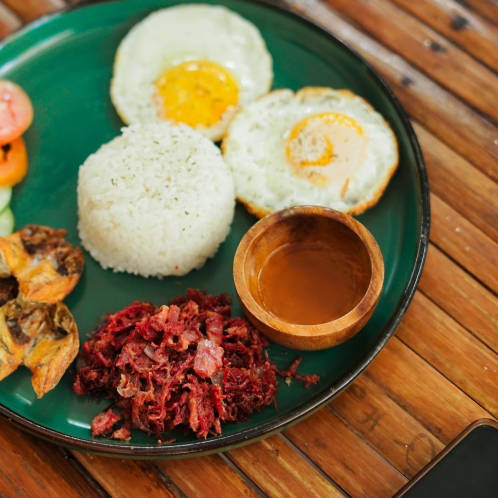 An overhead shot of a traditional Filipino boodle fight, featuring a variety of dishes like fried fish, noodles, fried chicken, sausages, and a central salad, all arranged on banana leaves.