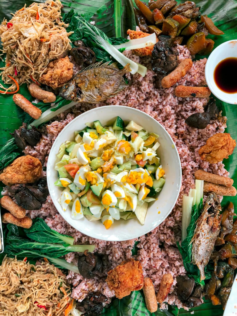 A close-up of a Filipino breakfast on a green plate, showing three sunny-side-up eggs, a mound of white rice, a serving of corned beef, dried fish, and a small wooden bowl of sauce, all on a bamboo mat.