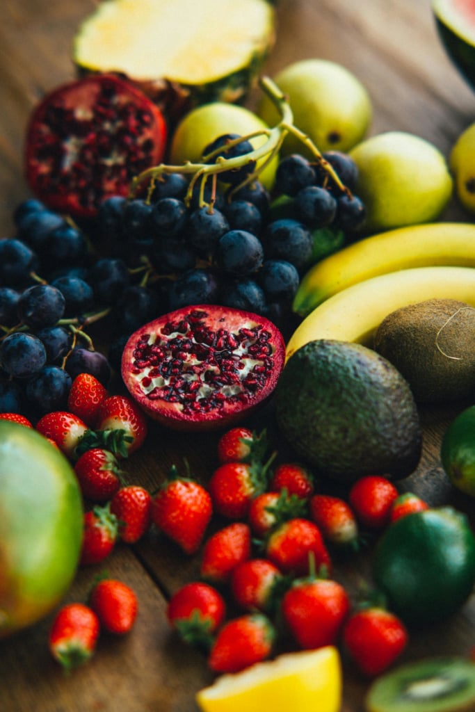  A colorful assortment of fresh fruits, including grapes, strawberries, pomegranates, bananas, kiwi, and an avocado, are artfully arranged on a wooden surface.