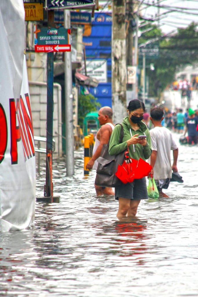  A street is flooded with waist-high water, and a person in a green hoodie and black mask stands in the water, looking at their phone and holding a bright red umbrella. Other people are wading through the floodwater behind them, and street signs and buildings are visible in the background.