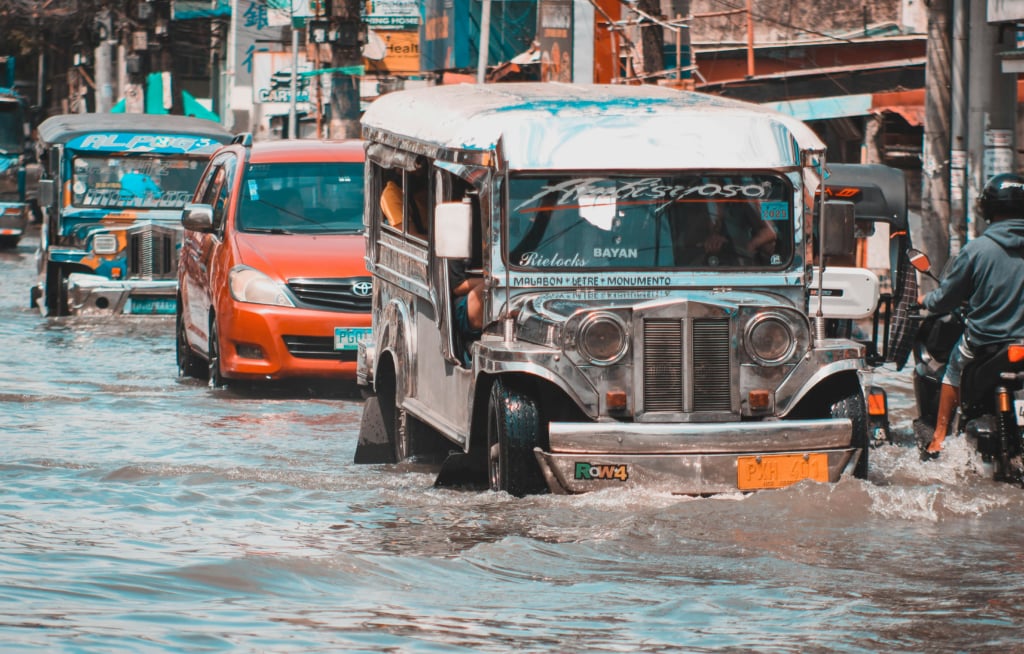  A street in Quezon City is flooded, with several vehicles, including a silver jeepney and a red car, driving through the murky water. A person on a motorcycle is on the right side of the frame, also navigating the flood.
