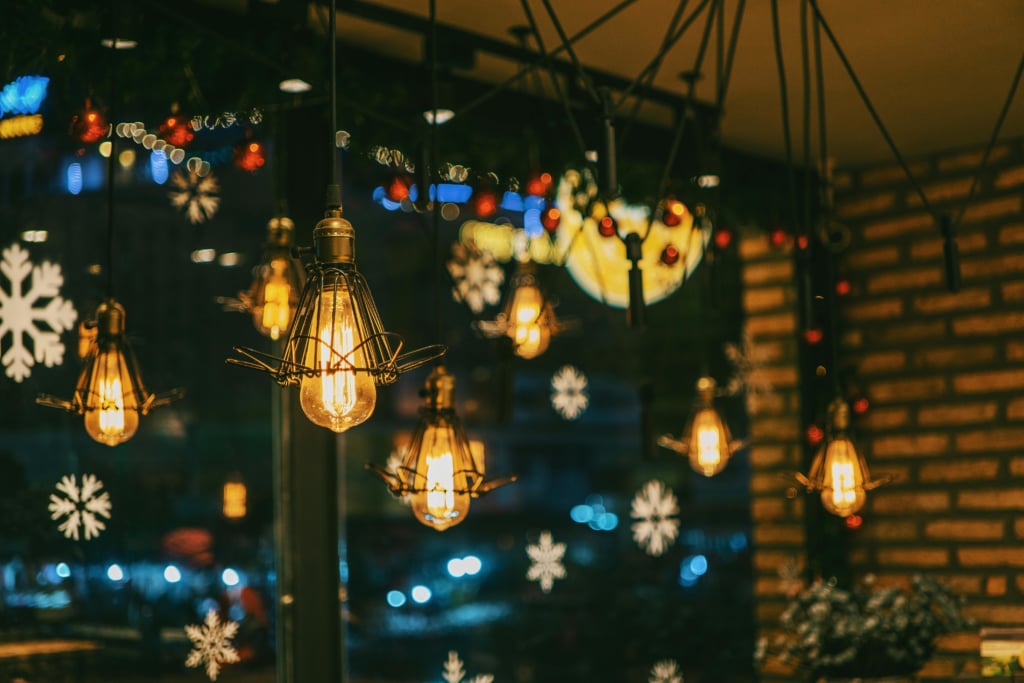  A warm, moody indoor shot featuring several vintage Edison-style light bulbs hanging from the ceiling. The background is a window looking out onto a dark street with holiday decorations visible, including strings of red and blue bokeh lights and white snowflake clings on the glass. The brick wall texture in the background suggests a cozy cafe or restaurant setting.