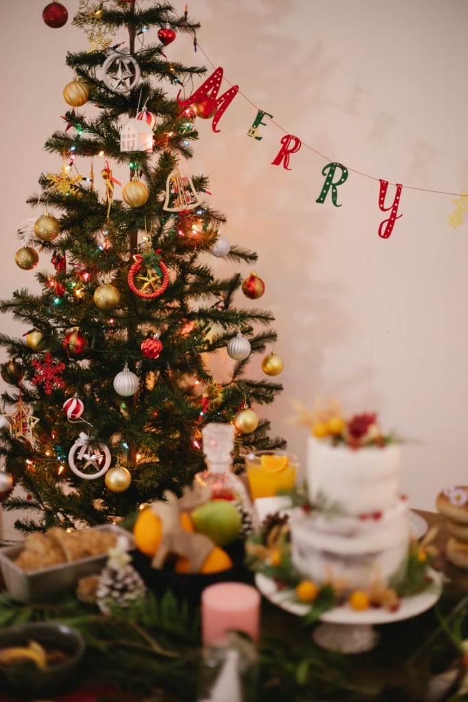  A slightly soft-focus vertical image of a festive indoor Christmas scene. On the left, a decorated Christmas tree is visible, adorned with lights, gold and silver ornaments, and various small decorations. 