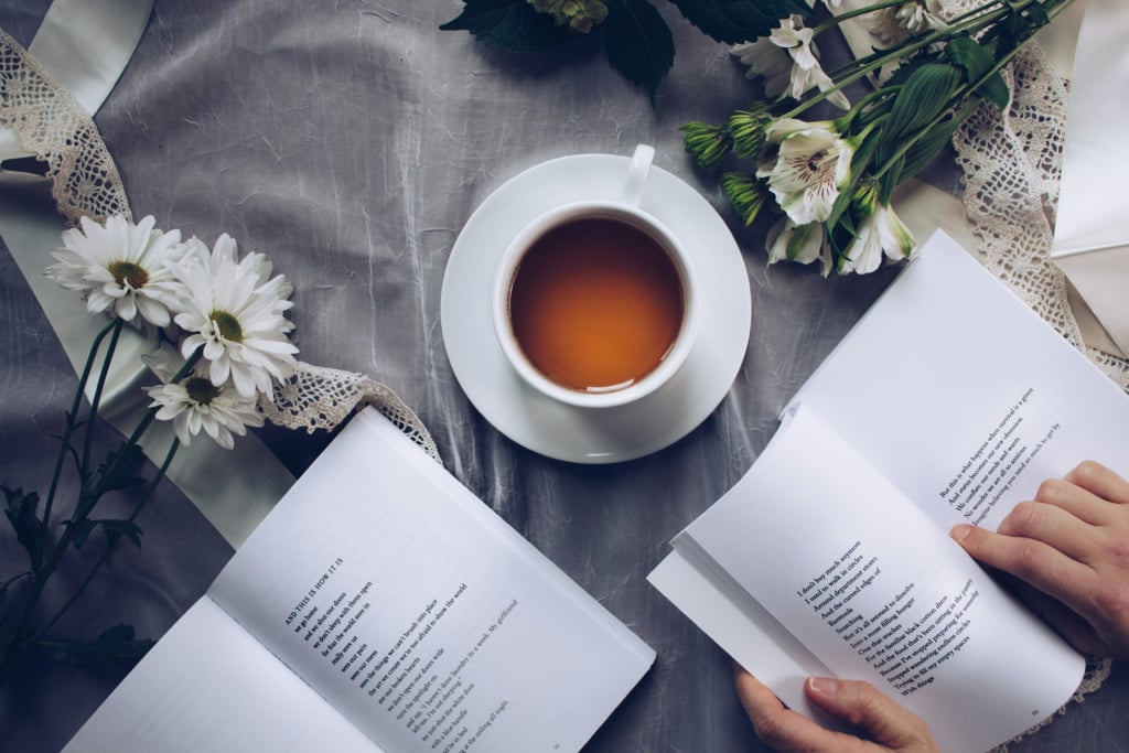 An overhead shot of two open books on a gray textured surface, with a white teacup and saucer centered between them. A hand gently holds the page of the book on the right. There are white daisies and other white flowers with green leaves decorating the top and left of the scene.