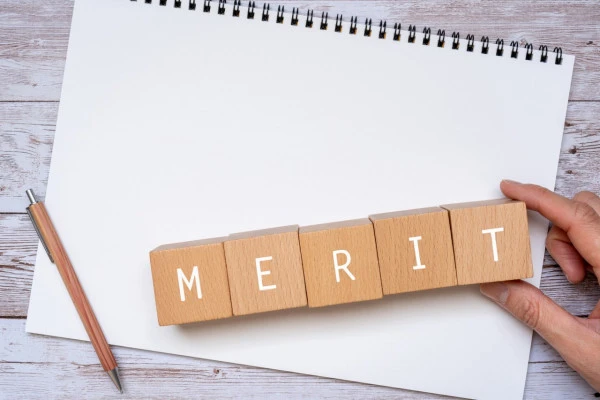 「MERIT」と書かれた積み木、ノート、ペン、人の手の画像