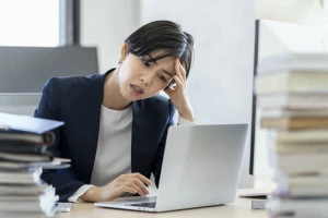 ノートパソコンの画面を見ながら頭を抱えるビジネスウーマンの写真