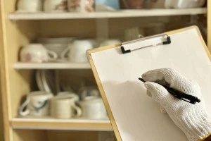 食器棚の前でクリップボードに記入する軍手をはめた手の写真