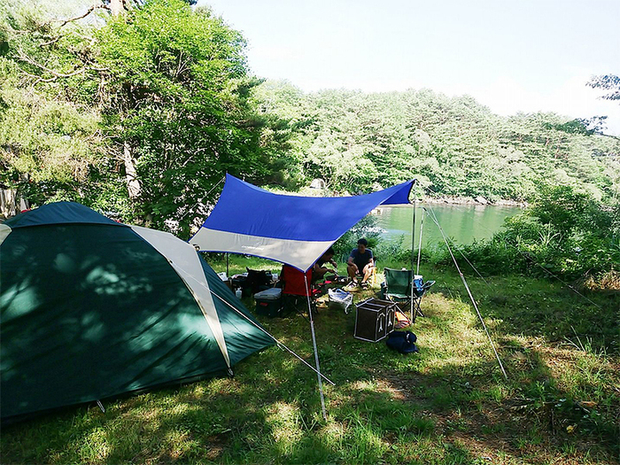 湖畔の林間で緑のドームテントに繋がる青いタープの下でくつろぐ2~3人