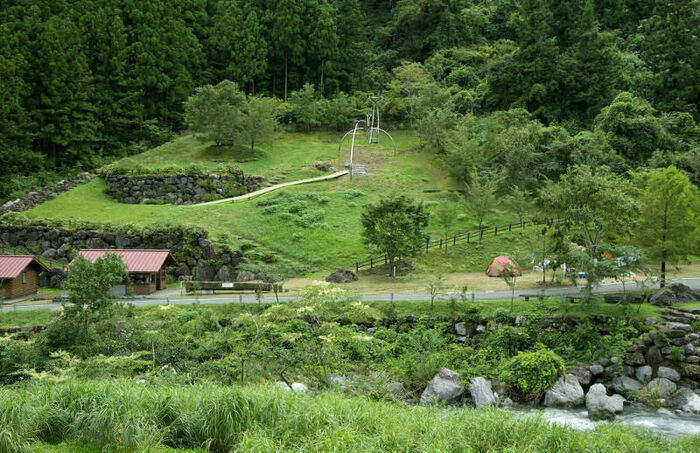 てまえに川が流れその奥に草地のキャンプサイトと山