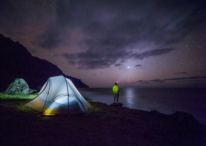 夜の湖の淵辺で湖を眺める男性とテント内の明かりを灯している石油ストーブ
