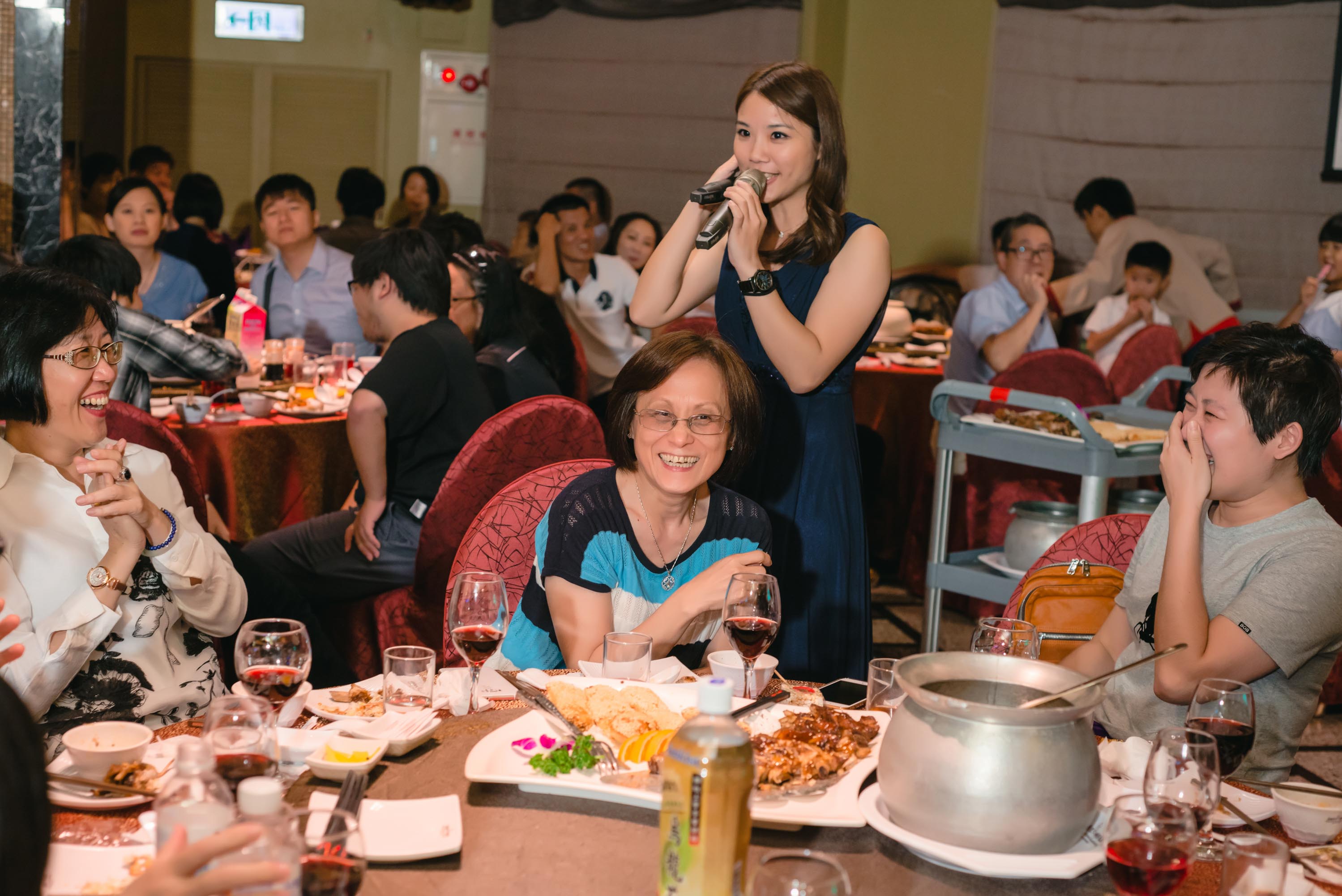 這張照片是一群人在餐廳用餐,其中一個穿著藍色洋裝的女生正拿著麥克風唱歌,旁邊還有一個人正在演奏吉他,氣氛看起來很熱鬧。