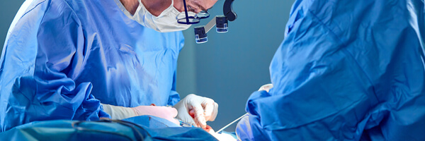 Surgeon and his assistant performing cosmetic surgery in hospital operating room. Surgeon in mask wearing loupes during medical procadure.