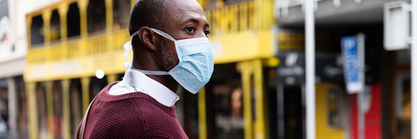 African American man out and about in the city streets during the day, wearing a face mask against air pollution and covid19 coronavirus, crossing the street and using his smartwatch.