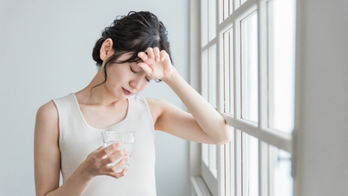 体調不良で頭に手を当てながら、窓辺で水の入ったグラスを持つしんどそうな女性のイメージ