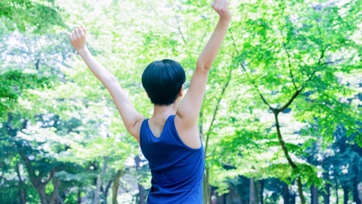青々とした鮮やかな新緑の木々が広がる森の中で、両腕を大きく上へ伸ばして気持ちよく深呼吸やストレッチをしている女性の後ろ姿。