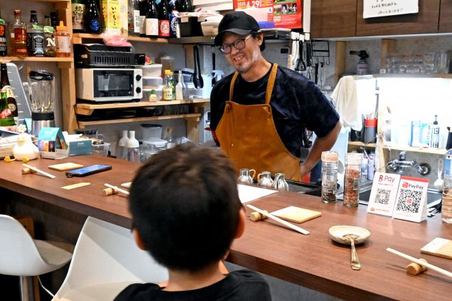 角打ちを切り盛りする松田健太郎さん。手前の男の子はクレープを食べにきた〝常連〟だそう。「角打ち」にもかかわらず、子どもがいるのはなぜかというと&hellip;＝2025年10月、大阪府豊中市、松尾葉奈撮影