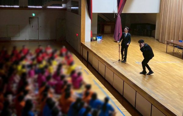 体育館のステージで生徒に漫才を披露する「キクバリ」＝キクバリ提供、画像の一部を加工しています