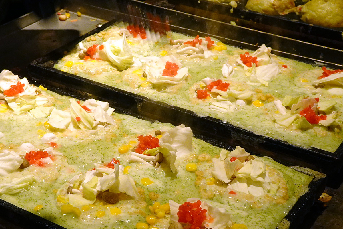 「爆彈燒本舖」（ばくだん焼本舗）秉持著「要吃，就得選最有料」的硬派職人精神，將「天婦羅碎、章魚、蛤蜊、玉米」等十多種嚴選食材濃縮進酥脆外皮中。