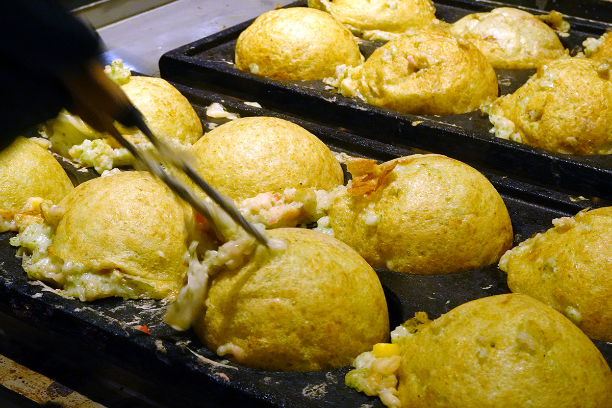 「爆彈章魚燒」（ばくだん焼）的核心靈魂在於其「厚實又豐盛」，每一顆都必須經過職人長時間的精準翻轉，才能維持外皮金黃酥脆、內餡炙熱又柔滑的日式口感。